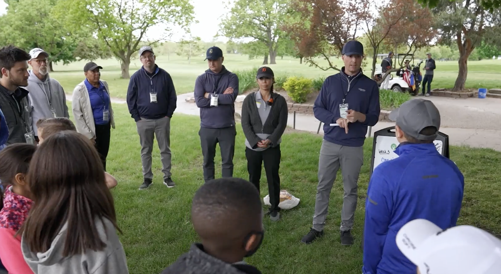 A group of people attending an outdoor golf course event or class, listening to an instructor
