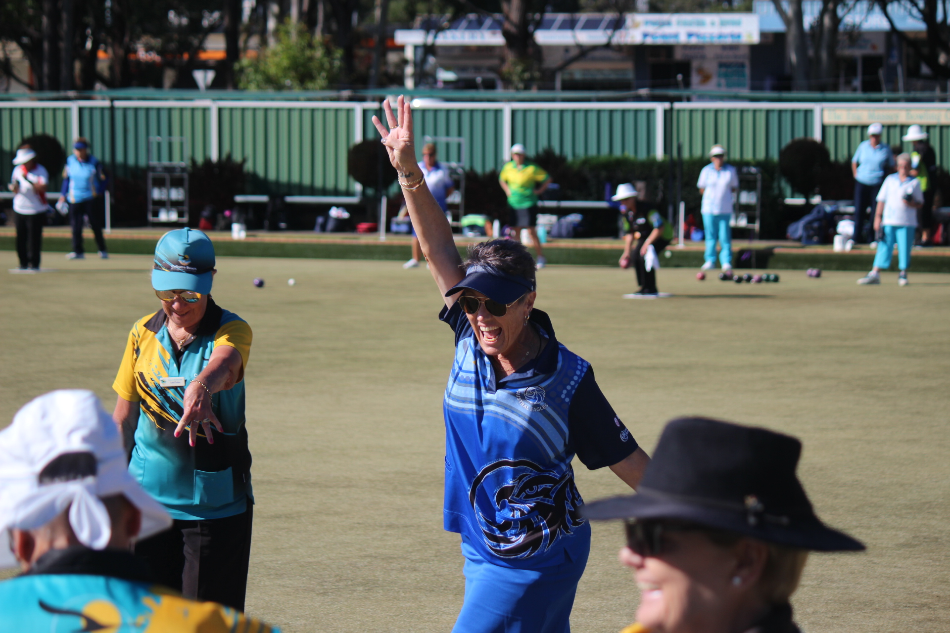 Woman lawn bowls player in a blue uniform and visor raises her arm in celebration on a sunny outdoor green, with other bowlers and bowls visible in the background
