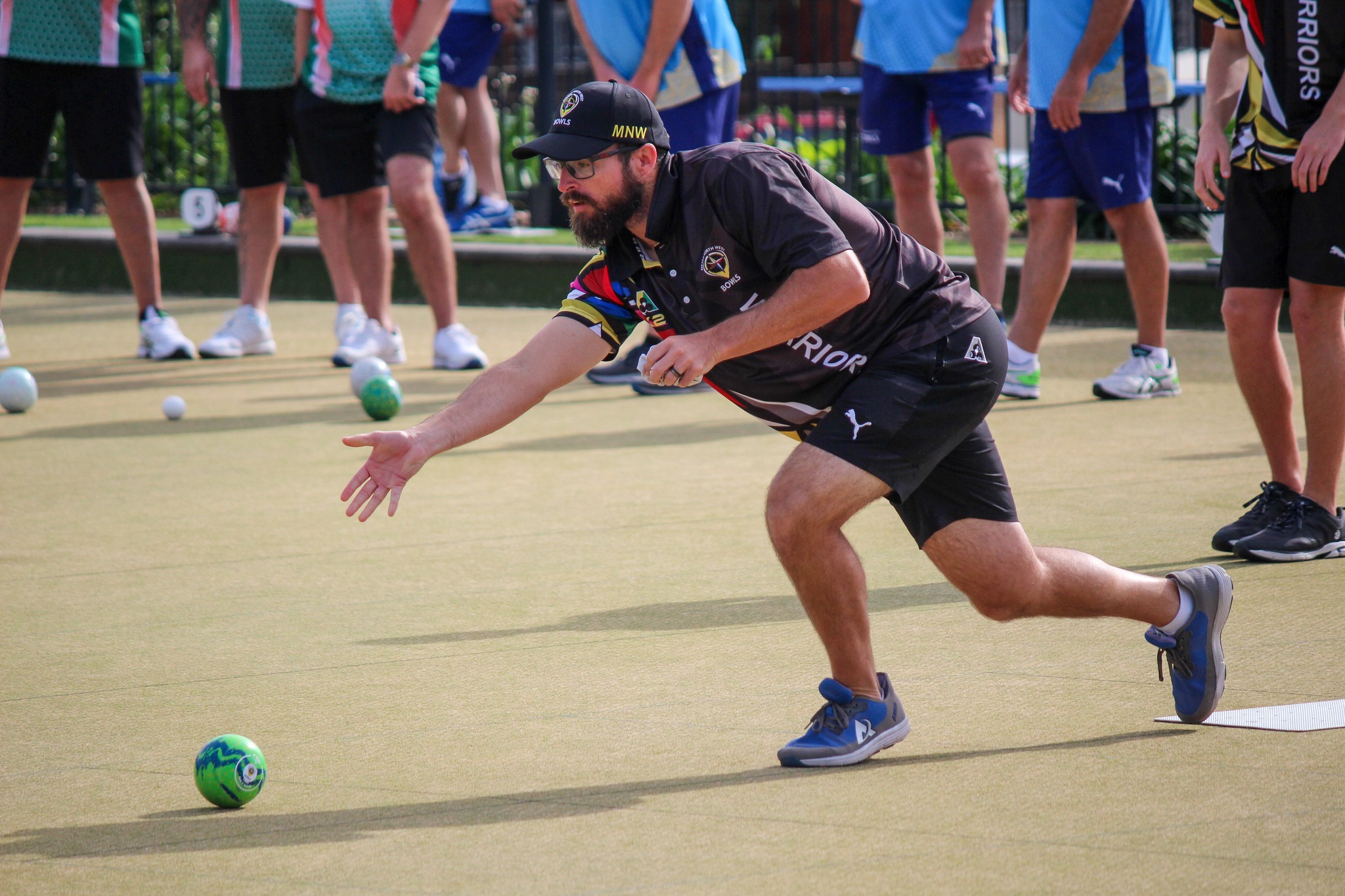 Lawn bowls player in a black team uniform and cap lunges forward to roll a green bowl on an outdoor green, with other players watching behind